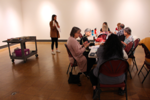 Image of a gallery space with two tables and a gallery cart. Jenna, a white woman with brown hair and tan colored coat and white sneakers, is holding a piece of paper and teaching a group of older individuals who appear to be engaged in an art-making activity. 